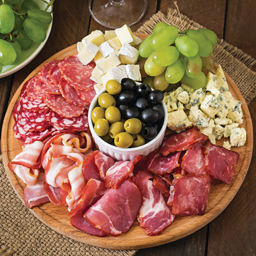 Wooden platter with cured meats, cheeses, and grapes on a woven mat.