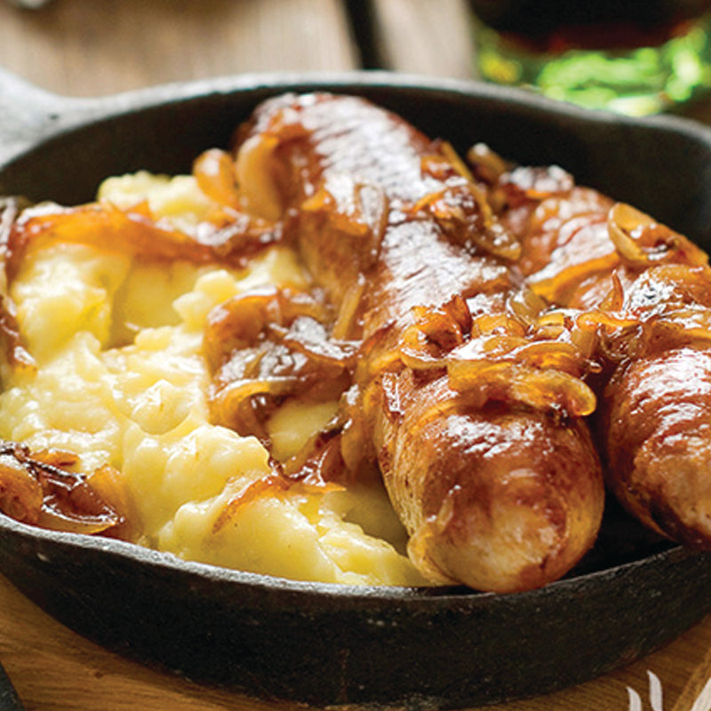 Two sausages with onions on a bed of mashed potatoes in a black skillet.