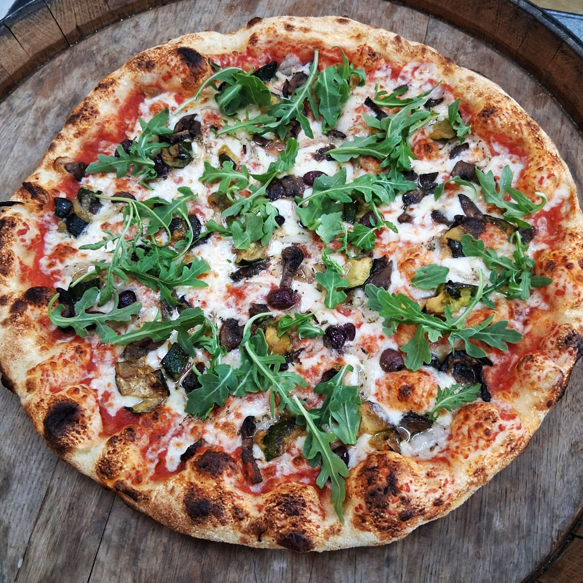 vegan pizza crafted with vibrant, quality ingredients. Tomato sauce pairs with creamy vegan mozzarella, while kalamata olives and sautéed mushrooms add depth. Caramelized onions bring subtle sweetness, fresh cherry tomatoes provide brightness, and peppery arugula finishes with a crisp, herbaceous note